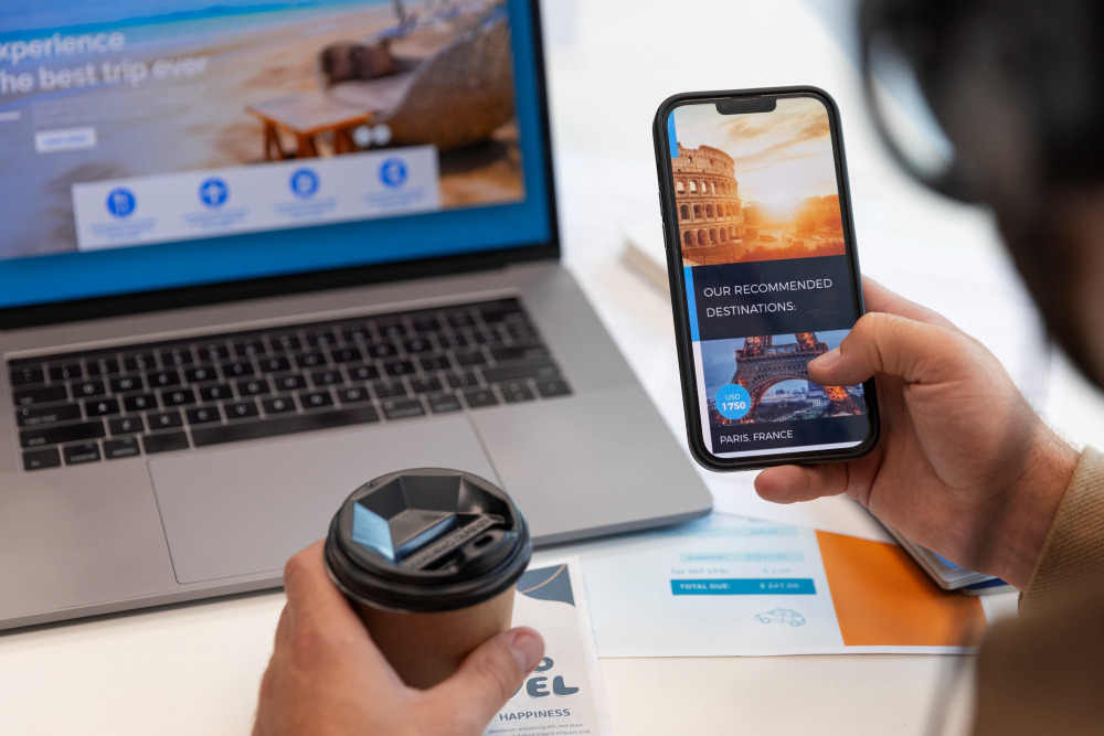 A man searches for travel websites on his laptop while using a smartphone for additional information